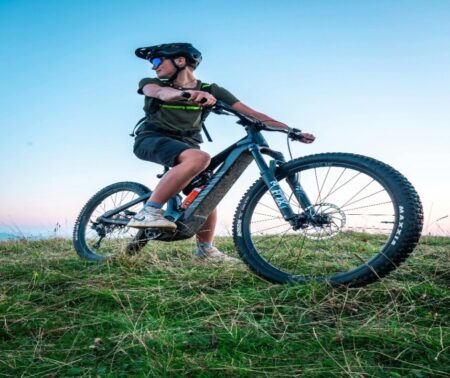 Un cycliste sur un vélo tout terrain, portant un casque et des lunettes de soleil, posant sur une colline herbeuse au coucher du soleil.